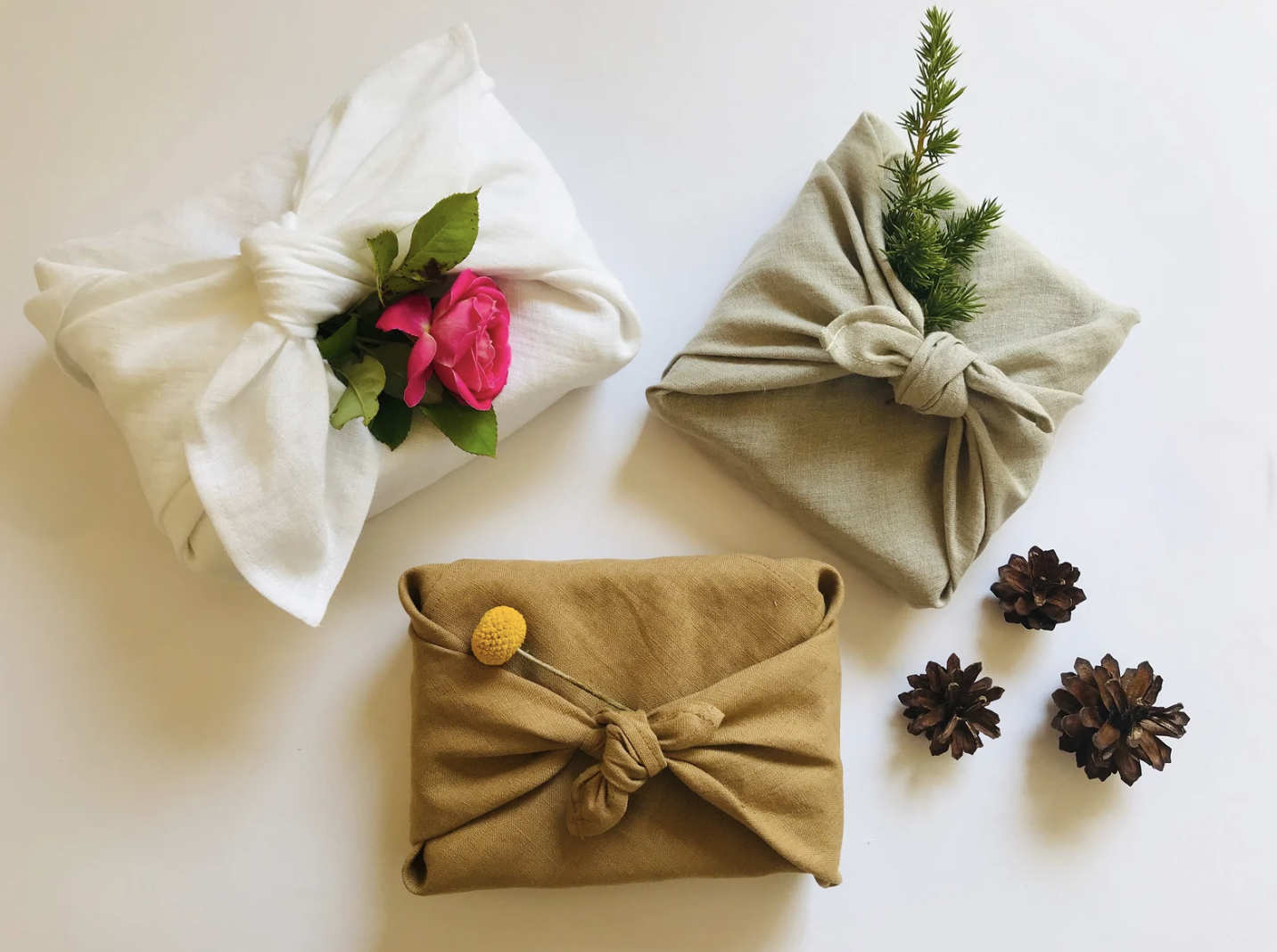  This image shows three gifts wrapped in fabric using the Japanese furoshiki method, tied with knots and adorned with natural elements like a pink flower, green sprigs, and a yellow billy button flower. Small pinecones are placed nearby on a clean white background, adding a natural and rustic touch