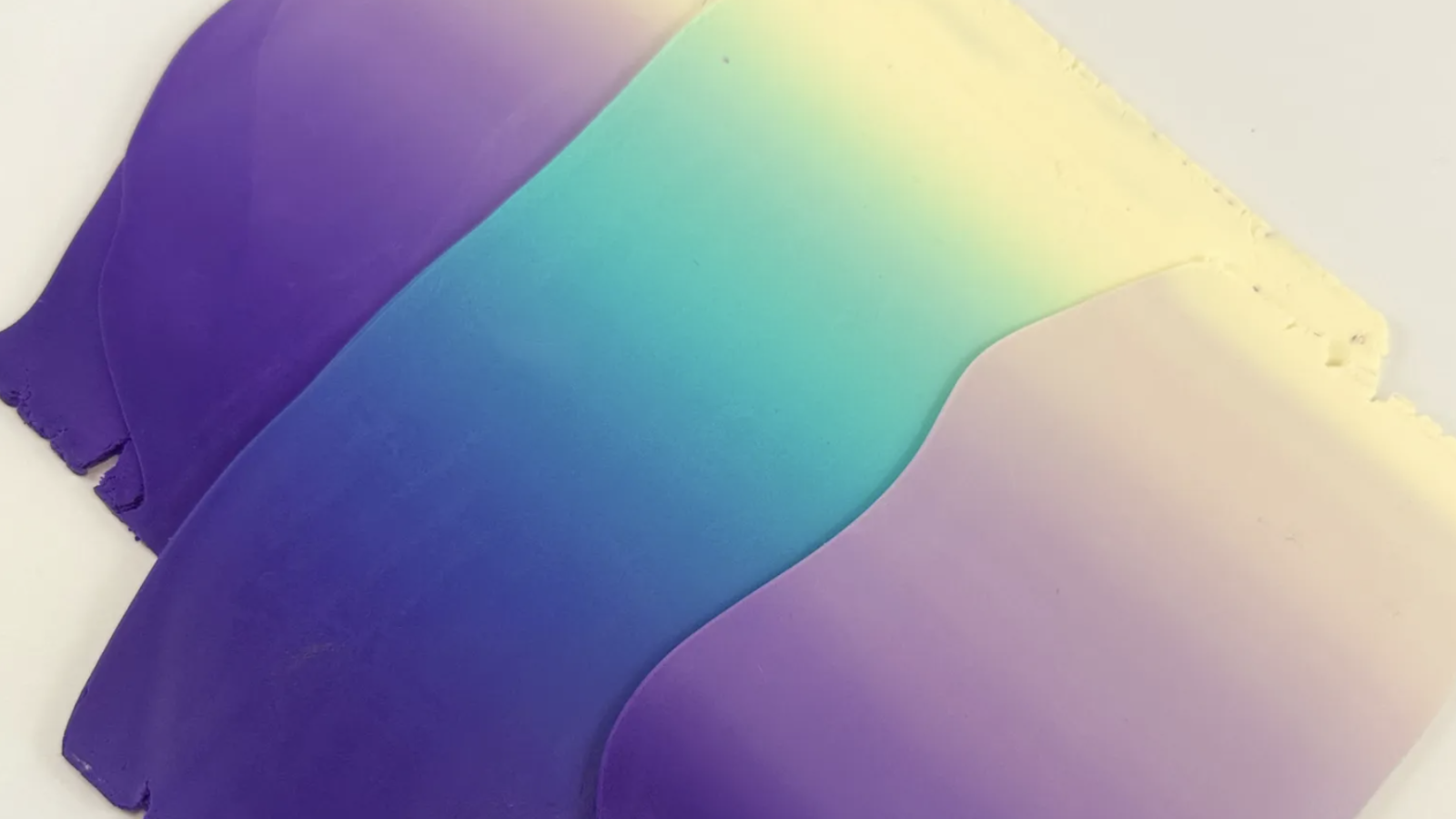 This image shows a smooth gradient of colors in polymer clay blending from deep purple to blue, teal, yellow, and cream, creating a visually striking ombre effect. The material appears layered or sculpted, with soft curves and slightly uneven edges, adding texture and depth.